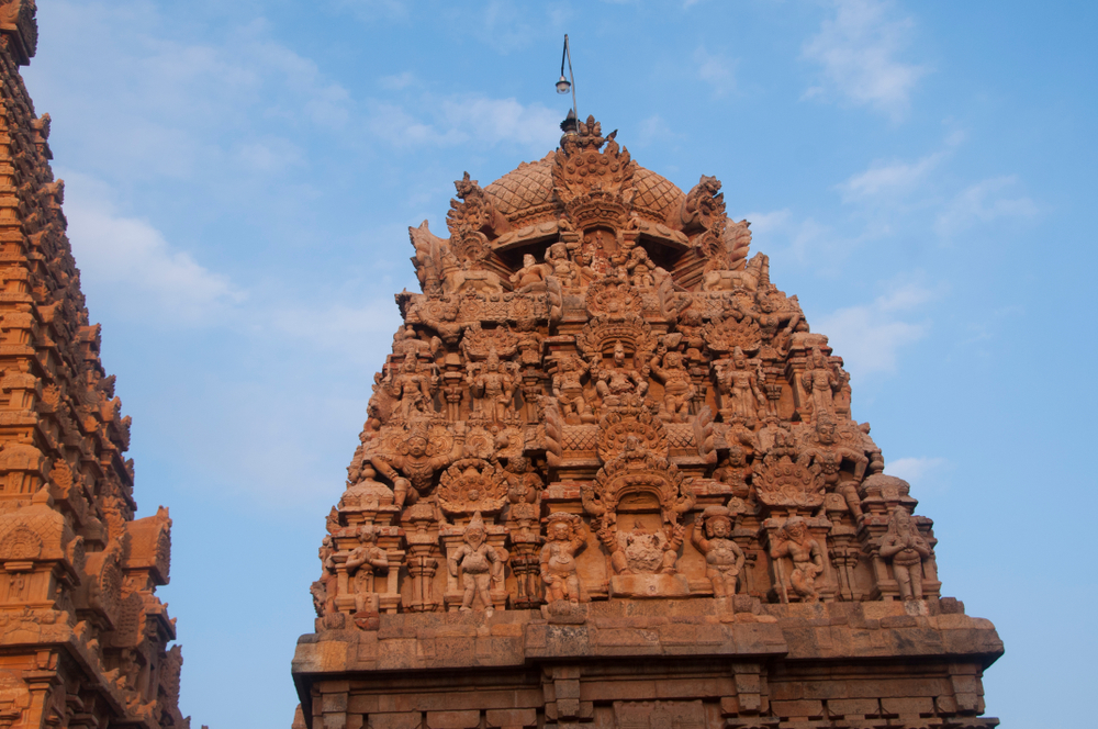 A neighbouring gopuram crown — densely populated with celestial figures — set against the planar vimana wall behind it